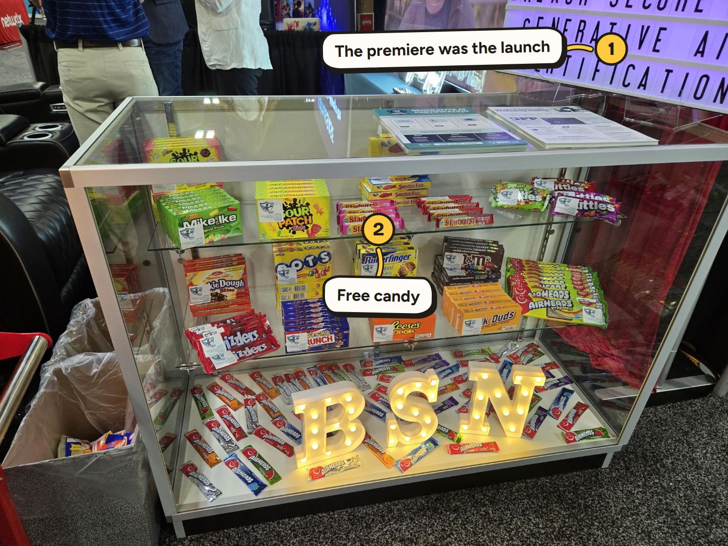 The movie-lounge booth at Pax8 Beyond 2025: a glass candy case of Breach Secure Now boxes above lit BSN letters. Marked on the photo: 1, The premiere was the launch; 2, Free candy.